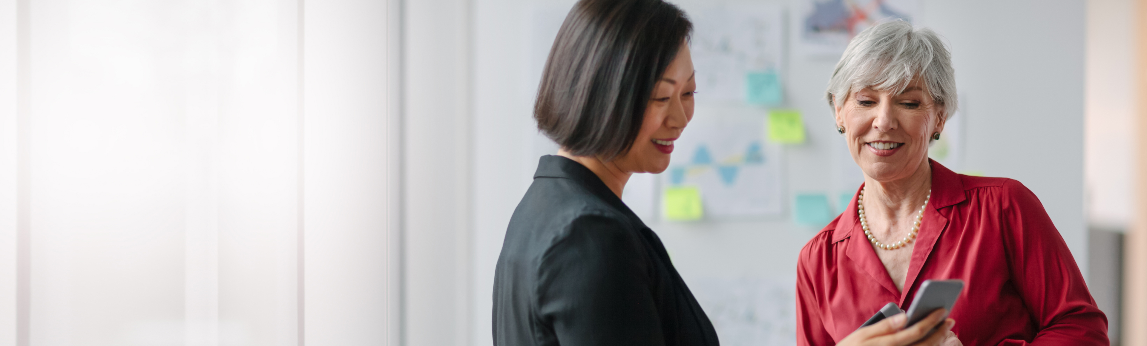 Caucasian senior businesswoman with Korean mature businesswoman standing and talking about ideas while looking at smartphone with charts diagrams well-dressed businesswear